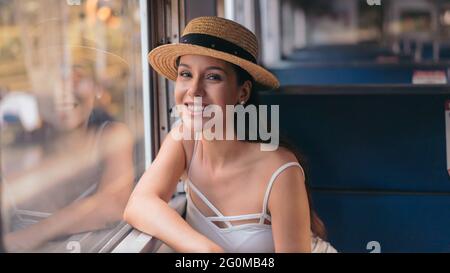 Portrait of beautiful young female tourist roaming around the city during vacation taking train as public transport while wearing a hat and smiling and looking at camera Stock Photo