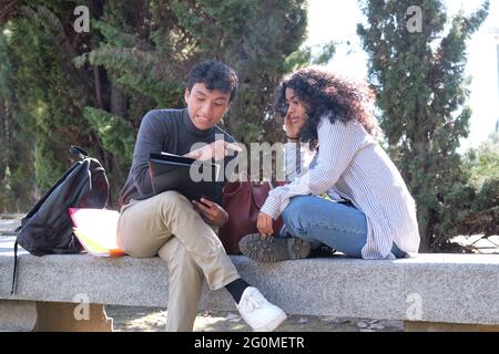 Two latin students studying together sitting on a bench outdoors. University life at campus. Stock Photo