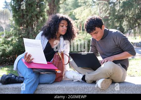 Two latin students studying together sitting on a bench outdoors. University life at campus. Stock Photo