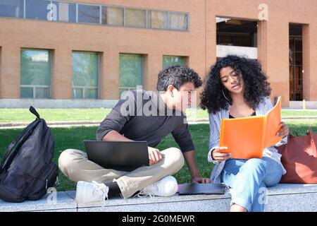 Two latin students studying from their lecture notes sitting on a wall outdoors. University life at campus. Stock Photo