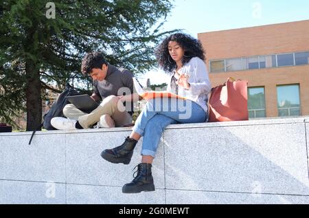 Two latin students studying from their lecture notes sitting on a wall outdoors. University life at campus. Stock Photo