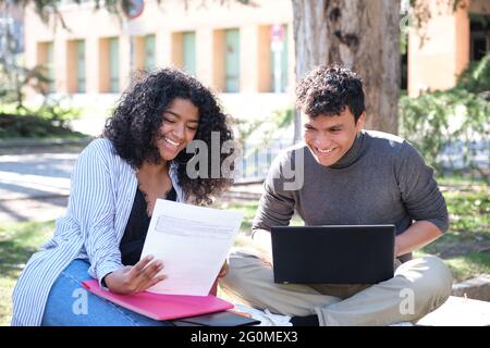 Two latin students laughing studying together sitting on a bench outdoors. University life at campus. Stock Photo