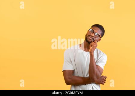 Good offer. Pensive black man in glasses looking up at copy space and touching his chin over yellow studio background Stock Photo