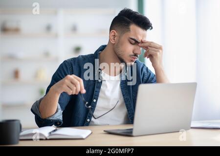 Exhausted arab guy sitting at workdesk, removing glasses Stock Photo