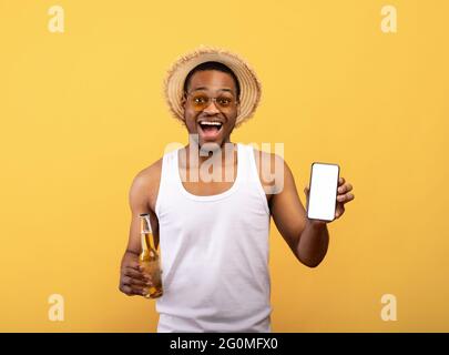 Funky black guy in summer clothes holding cellphone with blank screen and bottle of beer on yellow background, mockup Stock Photo