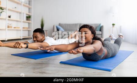 Black woman doing pilates workout using elastic strap sitting on yoga mat,  pulling training arms and shoulders in home living room. Athletic fit  exercising body using resistance band. Stock Photo