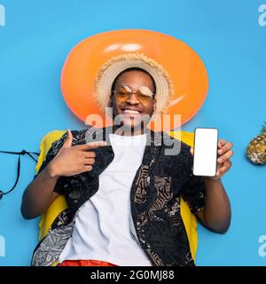 Handsome black guy in summer wear sunbathing on inflatable lilo, pointing at smartphone with white empty screen, mockup Stock Photo