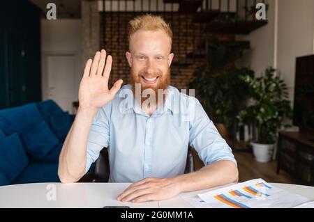 Smiling red-haired man looks at the camera and waving hand, video chat with with bearded hipster guy in smart casual shirt Stock Photo