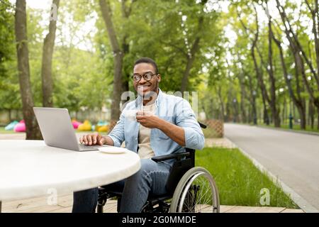 Happy handicapped black man in wheelchair using laptop for online job at cafe in city park Stock Photo