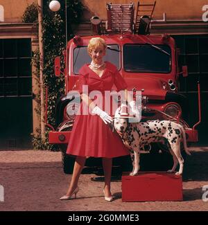 Fashionable in the 1950s. A young woman in a red dress, white gloves and white shoes is standing in front of a Ford firetruck together with a dalmatian dog with an american fire chief helmet. The dalmatier is traditionally a mascot of the fire brigade. Sweden 1958 ref CV68-2 Stock Photo