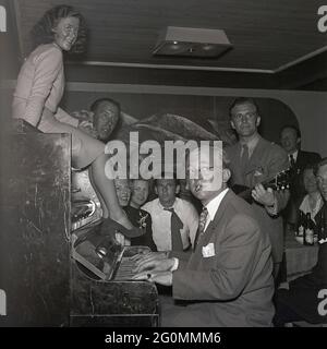Party in the 1940s. Two men are playing the piano but it looks as if it's only one, with four hands... Men and women are sitting around with drinks listening to the music,  a woman on top of the piano. The year is 1944, under the period of World War II. Sweden ref K9-4 Stock Photo