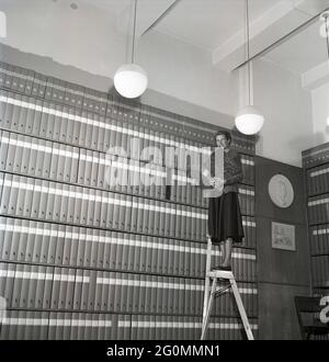 1950s woman at work. A young woman is standing on a ladder in an archive with shelfes filled with folders containing documents. Sweden 1953 ref BM25-3 Stock Photo