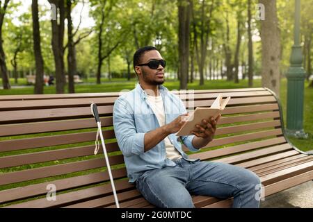 Young black visually impaired man sitting on bench in city park, reading Braille book outdoors Stock Photo