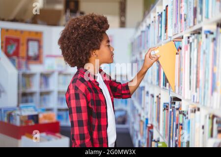 Happy african american schoolboy taking book from shelf in school library Stock Photo