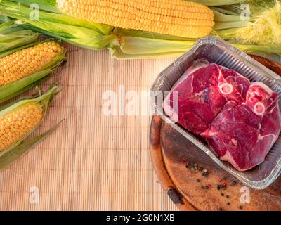 Flat lay of yellow corn cobs with green leaves on a table and in big pot and raw beef steak in a foil pan and whole pepper on a wooden cutting board. Stock Photo