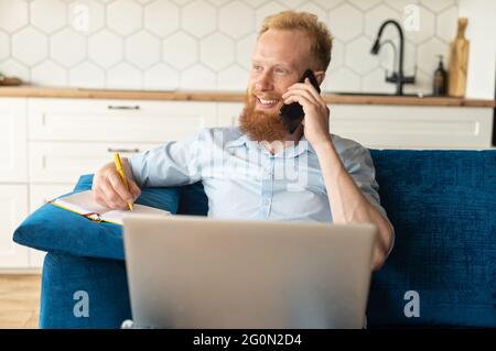 Cheerful redhead bearded male entrepreneur sitting on the sofa with a laptop on his laps, man talking on the smartphone and taking notes, making schedule Stock Photo