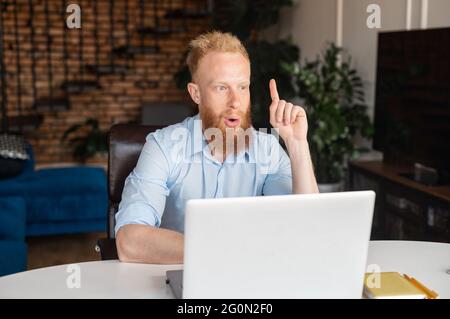 Redhead bearded man using computer laptop in the home office surprised with a new idea, points index finger up with amazed face expression, came up with a new task solution Stock Photo