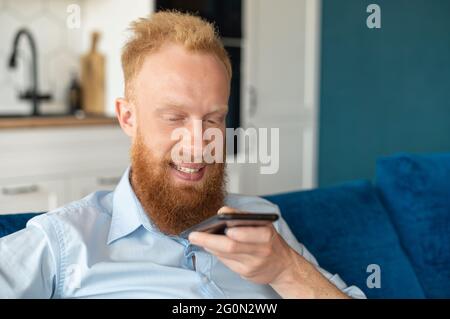 Charismatic redhead guy record voice message and smiling, sits on the sofa at home and holding smartphone near mouth and talking in the mic, chatting, using voice recognizable mobile application Stock Photo