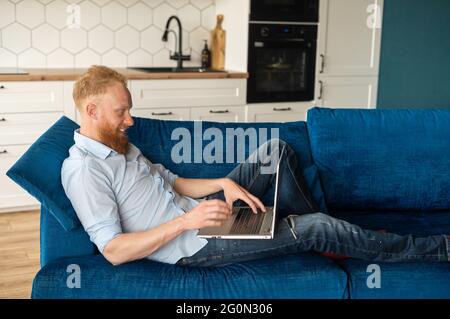 Handsome redhead bearded man lying down on the sofa and using computer laptop, smiling guy relaxing at home, browsing in networks, side view Stock Photo