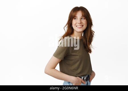 Modern happy girl with natural smile, standing in profile, look behind her shoulder with happy face, reading promotional text aside on white empty Stock Photo