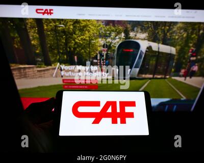 Person holding smartphone with logo of Construcciones y Auxiliar de Ferrocarriles S.A. (CAF) on screen in front of website. Focus on phone display. Stock Photo