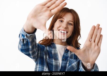 Close up of smiling redhead girl with freckles and white smile, look through hand frames gesture, taking shot of moment, searching for perfect angle Stock Photo