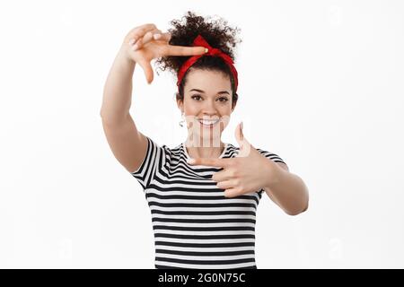 Portrait of creative young woman in headband, picturing moment, looking through hand frames gesture and smiling satisfied, searching perfect angle Stock Photo