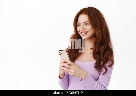 Portrait of lovely ginger girl chatting on smartphone, looking aside at someone with happy friendly smile, using mobile phone app, online shopping Stock Photo