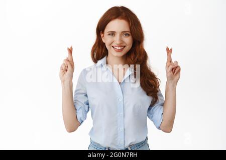 Portrait of optimistic redhead businesswoman cross fingers for good luck, look with confident, making wish, praying, waiting for results, standing Stock Photo