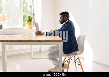 Side view of pensive African American man typing, taking notes, using laptop, holding online meeting with colleagues, involved in online conference Stock Photo