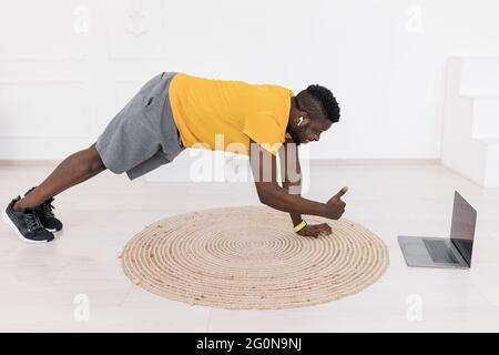 Happy young sporty man look at camera showing thumb up, while doing planck exercise,practicing work out using webcam lesson on laptop. Joyful active Stock Photo