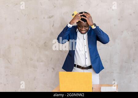 Stressed young Afro American male businessman, freelancer, or student stay at table by laptop smartphone overloaded by information from laptop Stock Photo