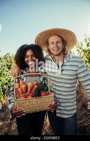 Mixed race farmers laughing and smiling after long day of harvest holding full basket of fresh vegetables  Stock Photo