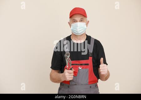repairman and plumber in protective mask giving a thumbs up, he is holding an adjustable wrench. Male worker with repair tool in overalls. Repair serv Stock Photo