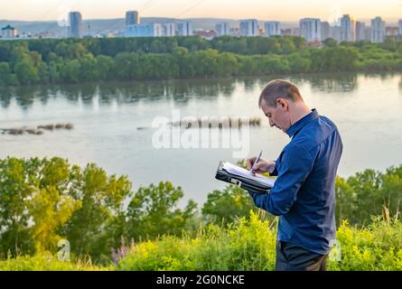 a man writes on a piece of paper while standing on a hill on the shore Stock Photo