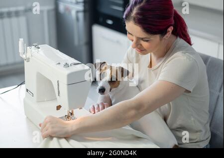 Caucasian woman sews while sitting in the kitchen. Dog Jack Russell Terrier sits on the lap of the owner. Home hobby. Stock Photo