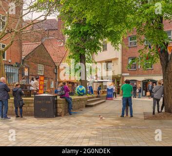 A tree overhangs a scene where a street performer attracts a small crowd.  Buildings and shops are in the background and people stand around. Stock Photo