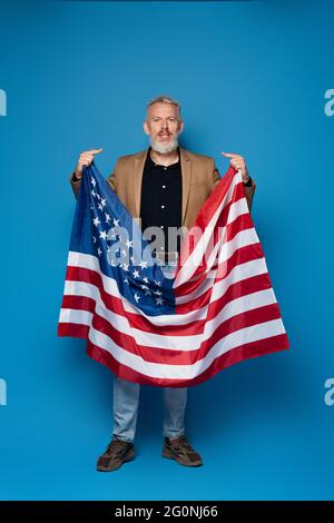 full length of bearded middle aged man standing with american flag and pointing at himself on blue Stock Photo