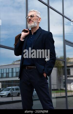tattooed mature man with grey hair holding leather bag while standing outside Stock Photo