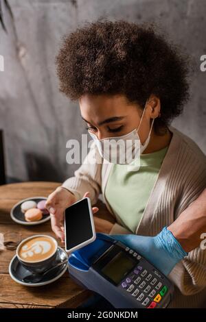 African american client in medical mask paying with smartphone near waiter with terminal in cafe Stock Photo