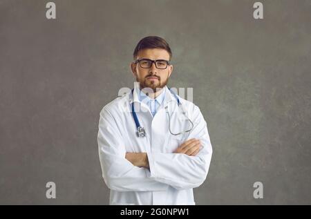 Focused serious male doctor in medical uniform stands with arms crossed on gray background. Stock Photo