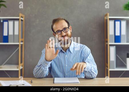 Happy office worker in glasses sitting at desk, smiling and waving hand at camera Stock Photo