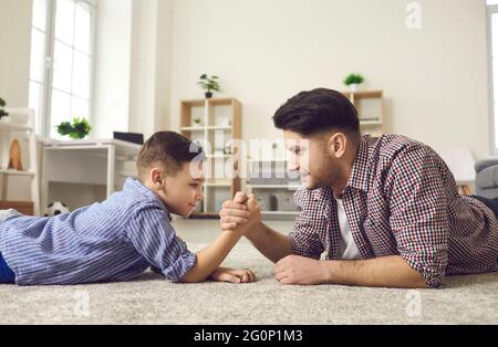 Side view of happy father and son competing in arm wrestling lying on the floor at home. Stock Photo