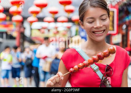 Chinese woman eating Bing Tang Hulu, a traditional chinese food snack from Beijing. Fruit candy stick sold in outdoor market place on Wangfujing Stock Photo