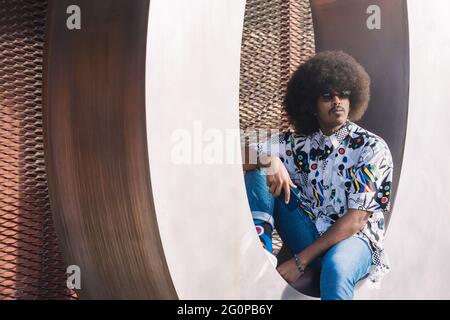 young guy with afro hair and sunglasses sitting on shiny metallic curved surface with red metallic grid in the background Stock Photo