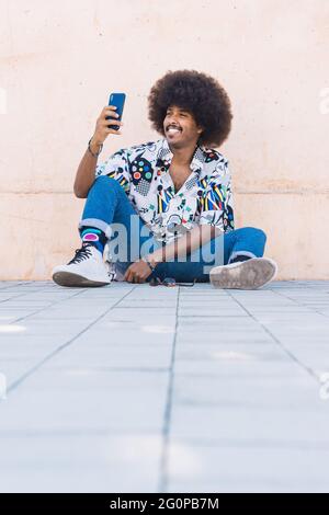 Smiling young black guy with afro hair taking a selfie sitting on the floor with unfocused foreground Stock Photo