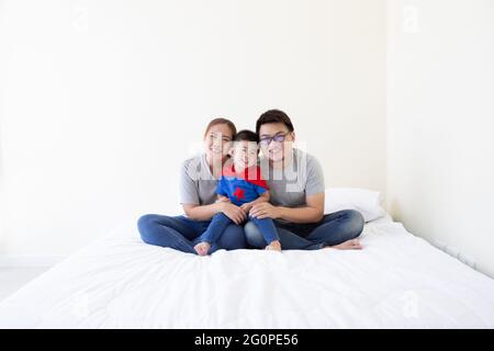Smiling Happy Asian family and son wearing superhero suit sitting on white bed in the bedroom Stock Photo