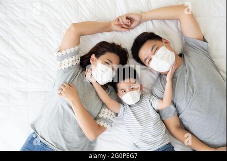 Top view of cute little boy and his asian parents looking at camera and wearing protective medical mask for prevent virus while lying on the white bed Stock Photo