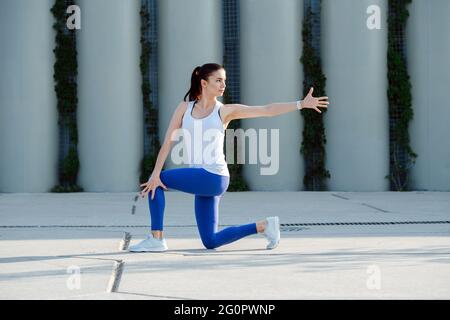 Young woman stretching on a concrete paving, doing twists, standing on one knee. Over support pillars. In blue stretchy yoga pants. Stock Photo