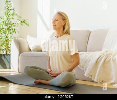 Peaceful senior woman in lotus position meditation with closed eyes at home while sitting on yoga mat on floor, full length. Calm elderly lady practicing meditation techniques and yoga indoors Stock Photo